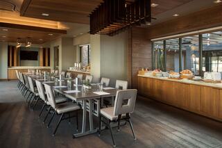 A modern dining space features a long table with cushioned chairs, large windows, and a buffet area with food options. Warm wooden accents enhance the ambiance.