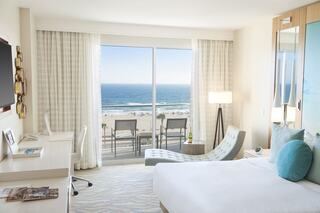 Bright, modern hotel room featuring a beach view, a cozy lounge chair, and a desk, creating a serene and inviting atmosphere.