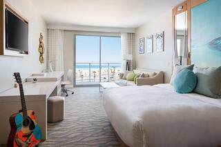 A bright, modern hotel room features a bed, couch, desk, and a scenic ocean view through large windows, with a guitar in the corner.