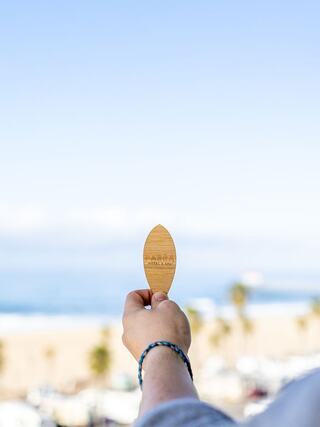 A hand holds a wooden object against a backdrop of a beach, ocean, and palm trees under a clear blue sky.