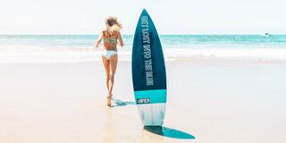 A woman in a bikini runs along a sandy beach, leaving a surfboard behind that reads "GET LOST INTO THE BLUE." Waves gently crash in the background.