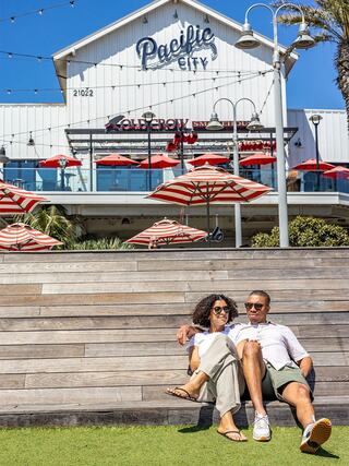 A couple relaxes on wooden steps amid vibrant umbrellas and palm trees, enjoying a sunny day at Pacific City.
