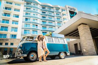 Two women pose beside a vintage blue Volkswagen van in front of a modern hotel with large windows and a stylish entrance. Sunlit and cheerful scene.