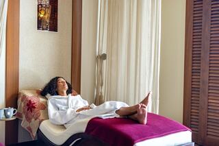 A woman relaxes on a lounge chair in a spa, dressed in a robe, enjoying a tranquil atmosphere with natural lighting and soft decor.