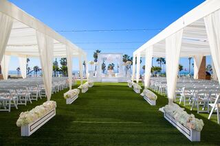 A beautiful outdoor wedding setup with white drapes, floral arrangements, and rows of chairs, overlooking a sunny beach and palm trees.