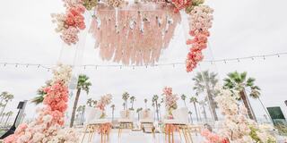 A beautiful wedding setup features a floral arch with pink and white blooms, elegant chairs, and palm trees in the background.