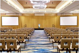 A spacious conference room with rows of beige chairs, flanked by large screens, and a decorative yellow curtain backdrop.