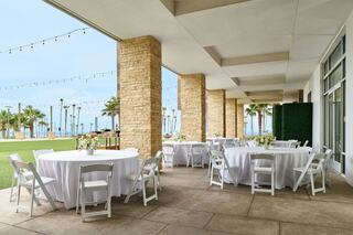 A spacious outdoor venue features round tables with white cloths, surrounded by palm trees and string lights, ideal for events.