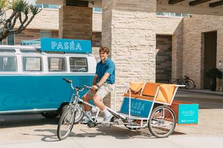 A young man in a blue shirt sits on a bike with an orange cart, next to a vintage van, promoting a hotel called PASÉA.