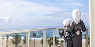 Two people in matching robes and towel wraps stand on a balcony, gazing out at the ocean and palm trees under a clear sky.