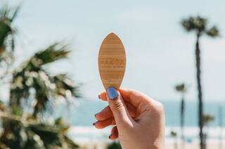 A hand holding a wooden paddle with a logo, set against a beach backdrop featuring palm trees and ocean waves.