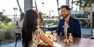 A man and a woman enjoy drinks at a stylish outdoor restaurant, smiling and engaging in conversation with a serene backdrop of palm trees.