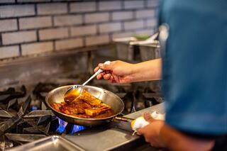 A chef expertly bastes cooking meat in a pan over a gas stove, surrounded by kitchen equipment and a brick wall.