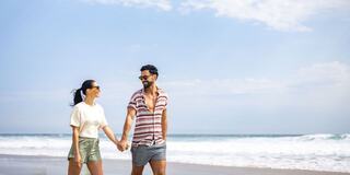 A smiling couple strolls hand in hand along a beach, with the ocean and a clear sky in the background, enjoying a sunny day together.