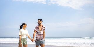 A smiling couple strolls hand in hand along a beach, with the ocean and a clear sky in the background, enjoying a sunny day together.