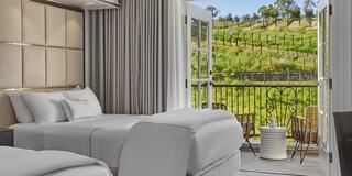 Stylish room with two beds, soft linens, and a balcony overlooking lush vineyards and green hills, creating a serene retreat.