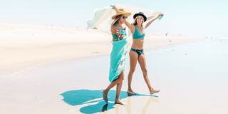 Two women walk along a sunny beach, dressed in swimsuits and beachwear, playfully enjoying the warm weather and water by the shoreline.