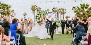 A joyful couple celebrates their wedding amidst palm trees, surrounded by enthusiastic guests, with elegant decorations and smiles all around.