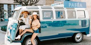 Two women in stylish outfits pose playfully by a vintage blue van with "PASÉA" signage, capturing a fun, sunny vibe.