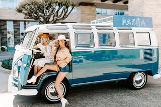 Two women pose playfully by a vintage blue van, dressed in stylish summer outfits with wide-brimmed hats, enjoying a sunny day.