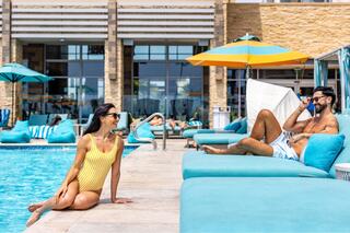 A woman in a yellow swimsuit leans by a pool, while a man relaxes nearby on a lounge chair, both enjoying a sunny day.