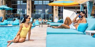 A woman in a yellow swimsuit sits by a pool, while a man in swimwear lounges, enjoying the sun and chatting on his phone.