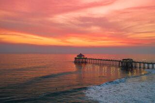 A vibrant sunset casts pink and orange hues over calm waters, with a pier extending into the sea, illuminated by soft lights.