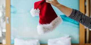 A hand holds up a festive red Santa hat with white fur trim against a soft blue background, conveying a cheerful holiday spirit.