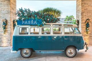 A vintage teal VW van with a "PASÉA" sign parked under palm trees, adorned with festive decorations and surrounded by stone walls.