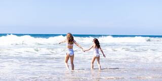 Two girls, one adult and one child, joyfully wade into the ocean, hand in hand, with waves splashing around them under a clear blue sky.