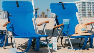 Two blue beach chairs sit on sandy shores, accompanied by a straw hat and sun care products, with palm trees and buildings in the background.