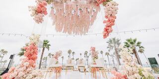 A romantic outdoor setup featuring an arch adorned with pink and white flowers, elegant chairs, and palm trees in the background. Perfect for celebrations.