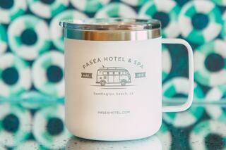 A white travel mug featuring the Pasea Hotel & Spa logo, depicting a retro van, set against a vibrant, patterned background.