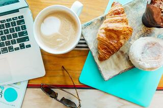 A laptop sits beside a cup of coffee and a plate with a croissant and pastry, complemented by sunglasses and a colorful notepad.