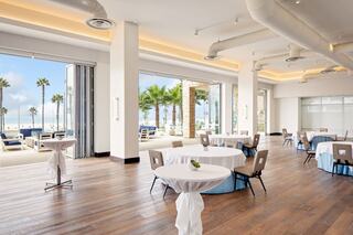 A spacious, modern beachfront venue with palm trees, large windows, and elegantly set tables, perfect for events or gatherings.