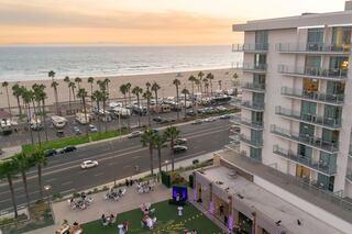 A lively beachfront scene at sunset, featuring palm trees, a busy street, and people enjoying their time on the grass and nearby beach.