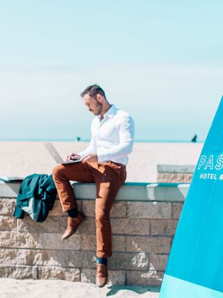 A man in a white shirt and brown pants sits on a beachside wall, working on a laptop with a surfboard beside him.