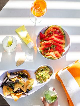 A vibrant display of fresh watermelon, guacamole, and nachos, paired with colorful cocktails and a cozy beach towel in sunlight.