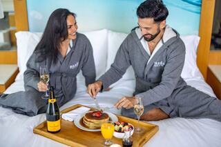 A couple in matching robes enjoys a cozy breakfast in bed, featuring pancakes, fresh fruit, and champagne. Laughter fills the air.