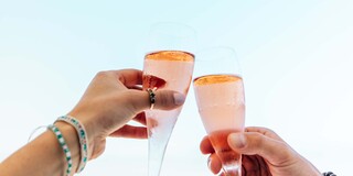 Two hands clink elegant champagne flutes filled with pink bubbly against a serene sky backdrop, celebrating a special moment.