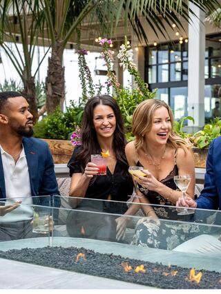 Four friends enjoy colorful cocktails at a stylish outdoor setting, surrounded by greenery and a cozy fire feature, sharing laughter and conversation.