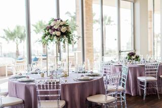Elegant tables with lavender linens and crystal chairs are adorned with floral centerpieces, set in a bright, open venue with palm trees outside.