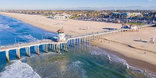 A long wooden pier stretches over gentle waves, with a sandy beach filled with visitors and palm trees lining the vibrant coastal area.