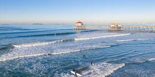 A serene beachfront scene features gentle waves, surfers enjoying the water, and a wooden pier with a vibrant red-roofed structure under clear skies.