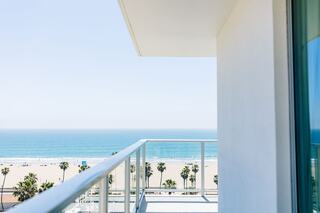 A clear blue sky meets the ocean, with a sandy beach lined by palm trees visible from a modern balcony.