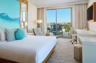 A modern hotel room featuring a large bed, light decor, a work desk, and a balcony with a scenic view. Bright and inviting atmosphere.