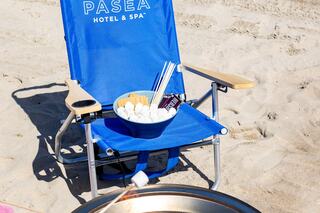 A blue beach chair sits on the sand beside a bowl of snacks, with straws and ingredients for a fun day by the shore.