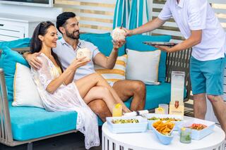 A couple enjoying drinks and food while lounging on a bright, cozy sofa outdoors, with a server bringing more refreshments.
