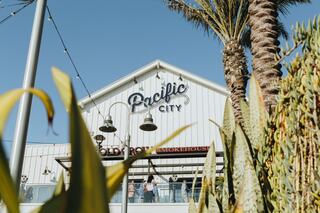 A vibrant coastal building with "Pacific City" signage, surrounded by palm trees and greenery, creating a welcoming atmosphere.