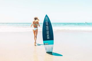 A young woman in a bikini walks towards the ocean, leaving a surfboard on the sandy beach, under a clear blue sky.